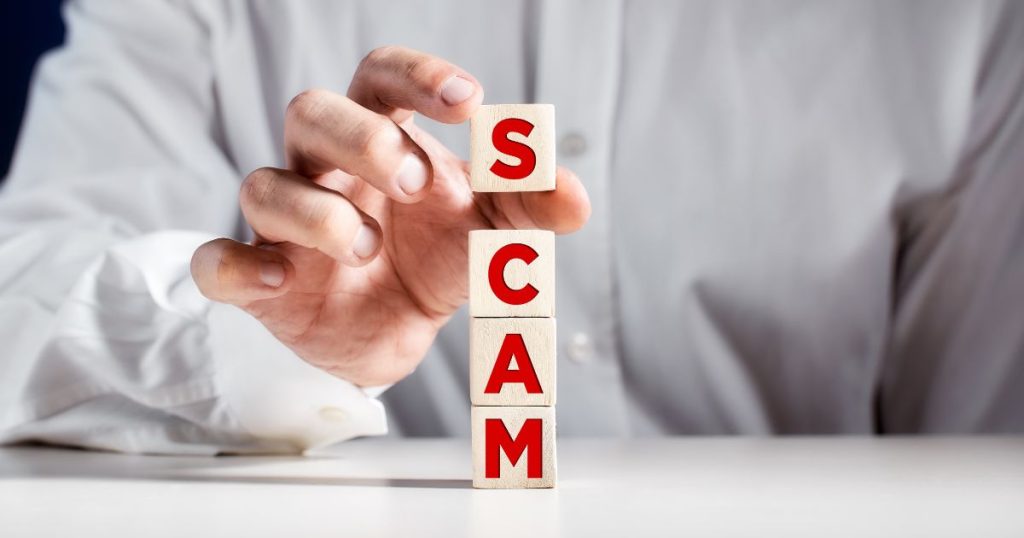 A person in a white shirt arranging wooden blocks that spell out the word "SCAM" in red letters, symbolizing the concept of a scam or fraudulent activity.