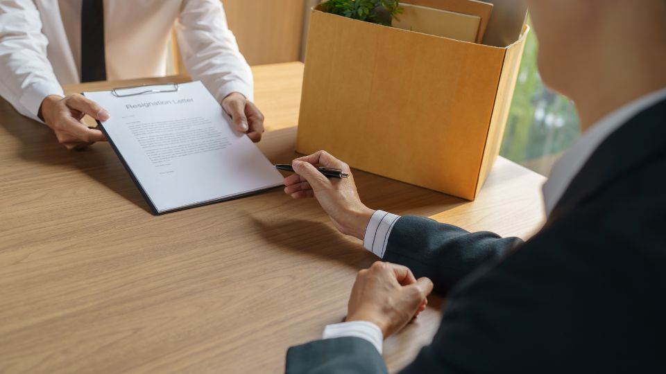 Man offering resignation letter to sign on, with a box of his things on the table 