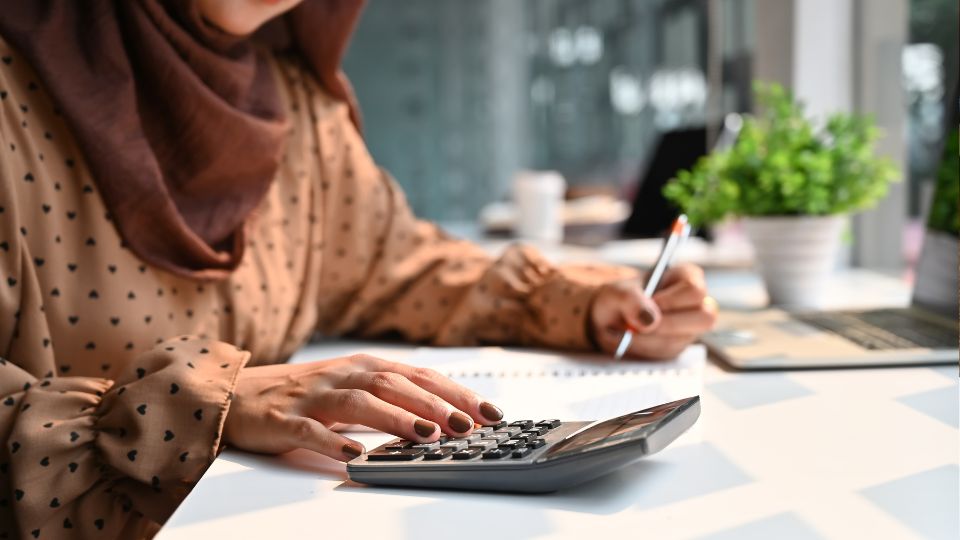 a hijabi Woman calculating something on the calculator and holding a pen 