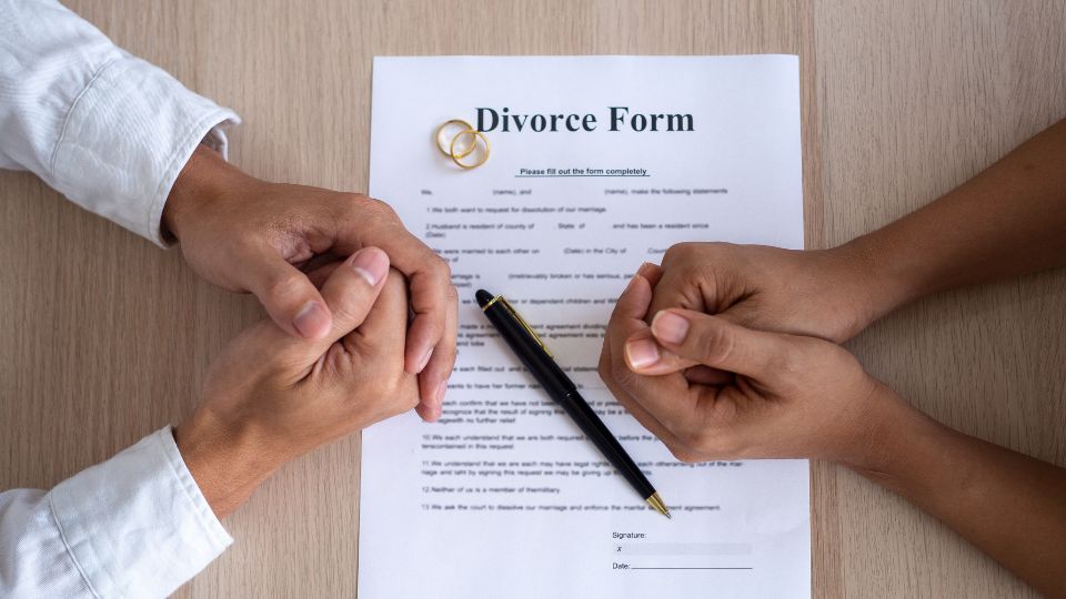 Two hands, one with a removed wedding ring, rest on a table near a divorce form with a pen, symbolizing the process of finalizing a marital separation.