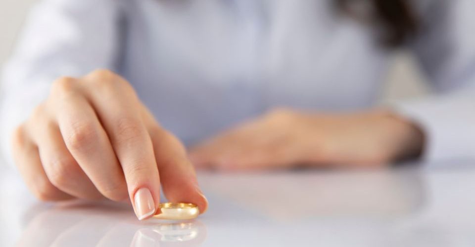 A person's hand with a manicured fingernail is poised to pick up a single golden-colored capsule from a white surface, suggesting the action of taking a supplement or medication.