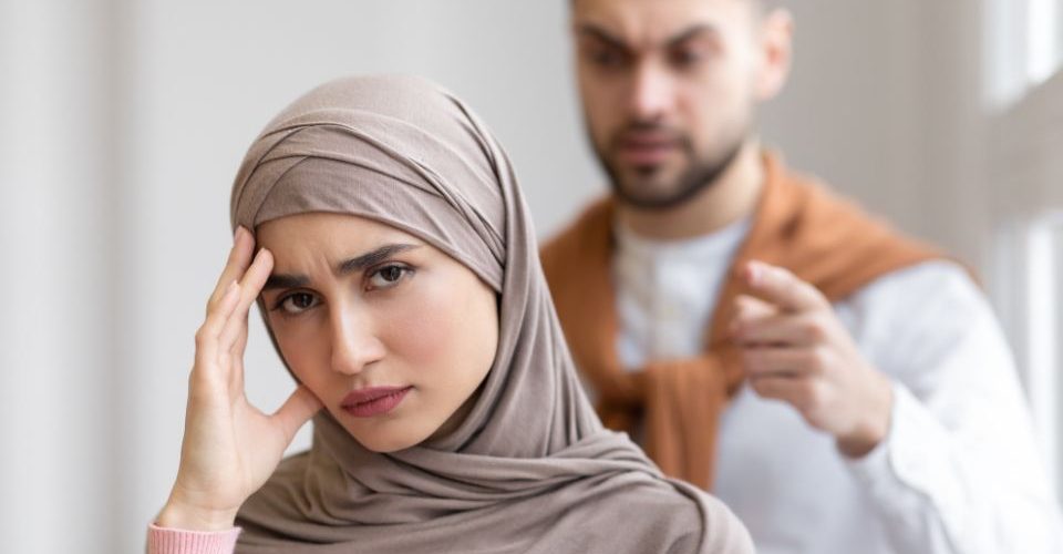 A woman wearing a hijab appears troubled or distressed while a man in the background gestures as if he is speaking to her in a serious or potentially argumentative manner.