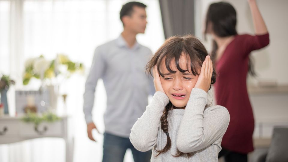 A distressed young girl covering her ears with her hands, crying, as two adults appear to argue in the background. The setting suggests a domestic situation where the child is upset by the confrontation between the adults.