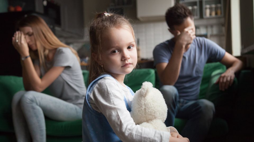 A young girl holding a teddy bear, looking solemnly at the camera, with a man and a woman sitting separately in the background on a couch, both appearing distressed and not facing each other, suggesting family conflict or emotional distress.