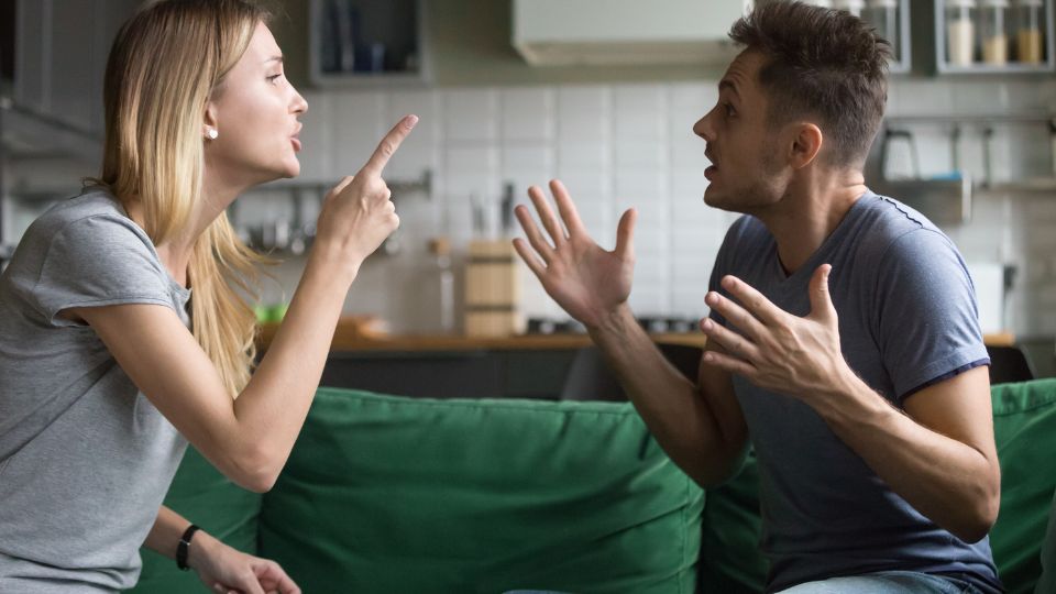 A man and a woman are engaged in a heated argument while sitting on a green couch, with expressive hand gestures and intense facial expressions that convey a sense of conflict or disagreement.