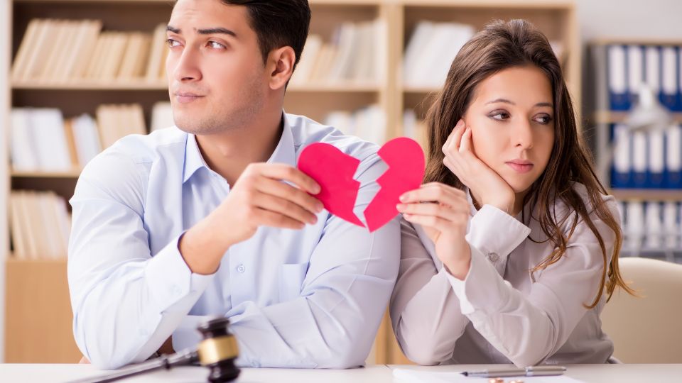 A man and woman sit at a table, with the man holding a torn red paper heart, signifying relationship troubles or the end of a partnership, while the woman appears contemplative or disappointed.