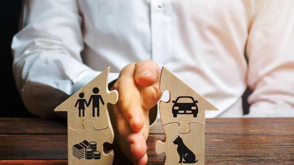 Two cardboard puzzle pieces in the shape of houses are being connected by a person in a white shirt. Each piece has different symbols: one with icons of two people and stacked coins, suggesting family and savings, and the other with a car and a dog, indicating vehicle ownership and a pet. This image metaphorically represents concepts such as family, home, finance, transportation, and pets as integral parts of life fitting together.