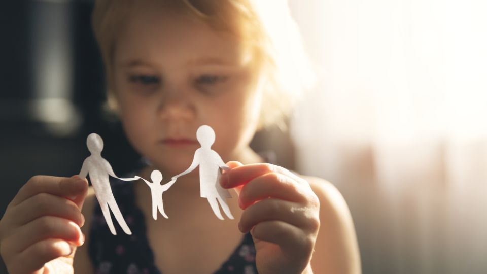 A young child holding two paper cutouts of figures representing adults and a smaller figure between them, with the figures on the ends being pulled apart. This image evokes the concept of a family being separated, symbolizing the impact of divorce or separation on children. The backlighting creates a somber and contemplative mood.