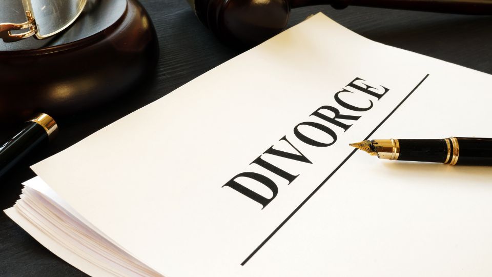 A close-up of a white paper with the word "DIVORCE" prominently printed on it, alongside a black fountain pen, suggesting the preparation for or the process of legal divorce proceedings. The dark surface and a vintage-looking gavel in the background contribute to the gravity of the situation.