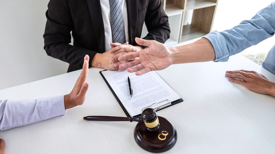 A formal setting where two individuals appear to be in a discussion or negotiation across a table, with a mediator or lawyer present. Legal documents and a gavel are visible on the table, indicating a legal context, and two wedding rings are prominently placed next to the gavel, suggesting the topic may be a divorce settlement or marital legal proceedings.