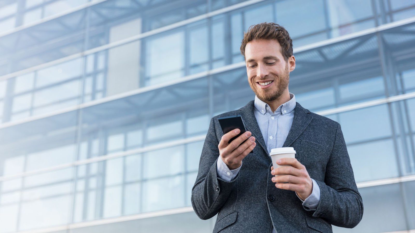 A smiling man in a smart casual outfit with a blazer and shirt, holding a mobile phone in one hand and a coffee cup in the other, standing in front of a modern glass building facade.