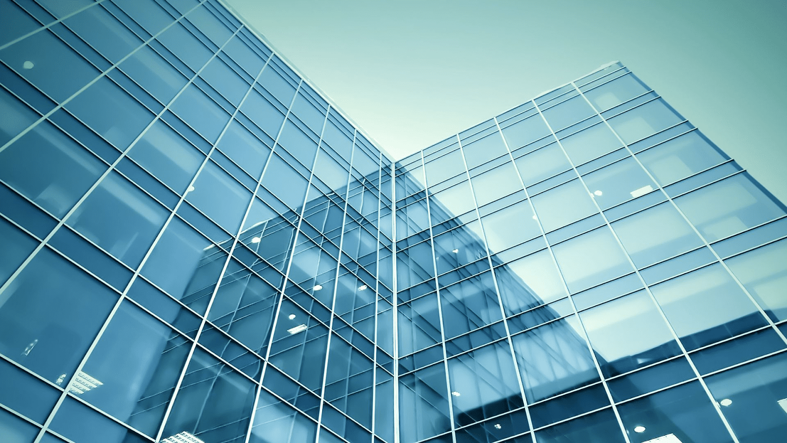 This image depicts the exterior facade of a modern building, characterized by its reflective glass panels that form a seamless curtain wall. The glass is tinted in a calming blue hue, which may indicate a design choice for aesthetic appeal or solar control. The structure's geometric precision, with its clean lines and right angles, suggests a contemporary architectural style, often associated with corporate or commercial buildings. The sky, barely visible as a reflection on the building's surface, is likely clear, hinting at the photo being taken on a bright day. This building could easily be part of a bustling business district, possibly housing offices such as law firms, financial institutions, or corporate headquarters.