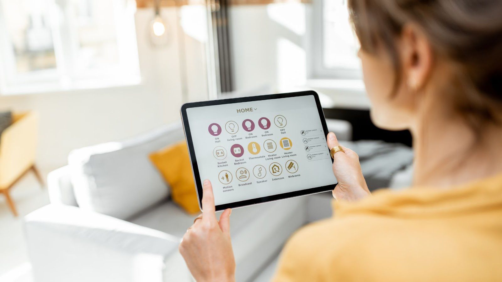 A person viewed from behind, focusing on a tablet displaying a smart home control panel with various icons for lighting, temperature, and security settings, in a bright living room setting.