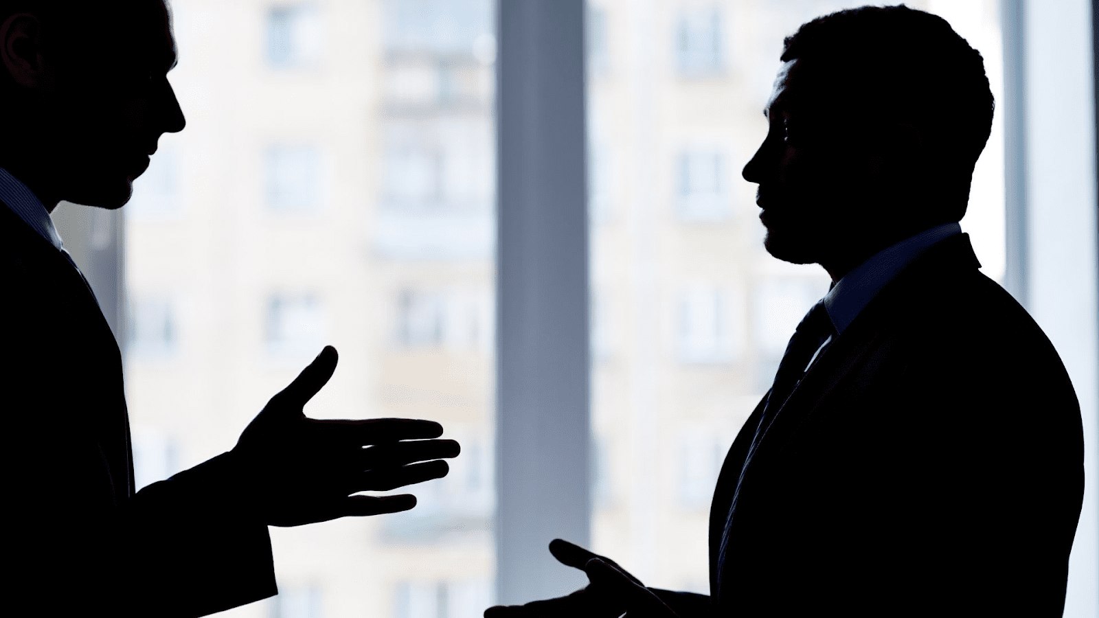 The image shows two silhouetted figures, presumably businessmen, in a discussion against the backdrop of a window with buildings visible outside. Their features are not discernible due to the backlighting, which creates a dramatic silhouette effect. One figure appears to be gesturing with their hand, indicating an active conversation or exchange of ideas. The context suggests a professional setting, possibly relating to corporate, legal, or financial matters.
