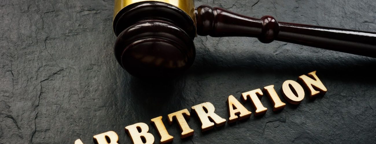 The image shows a wooden gavel, often used by a judge, resting on a dark surface with the word "ARBITRATION" spelled out in bold, capital letters, likely made of wood or metal, positioned in front of it. The composition suggests a legal context, specifically focusing on arbitration, which is a form of alternative dispute resolution outside of courts.