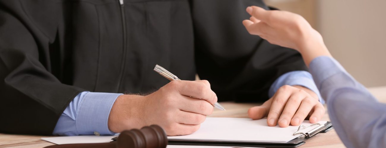 A person in a judge's robe writing on a document at a desk, with a client explaining a point, and a gavel in the foreground indicating a legal setting.