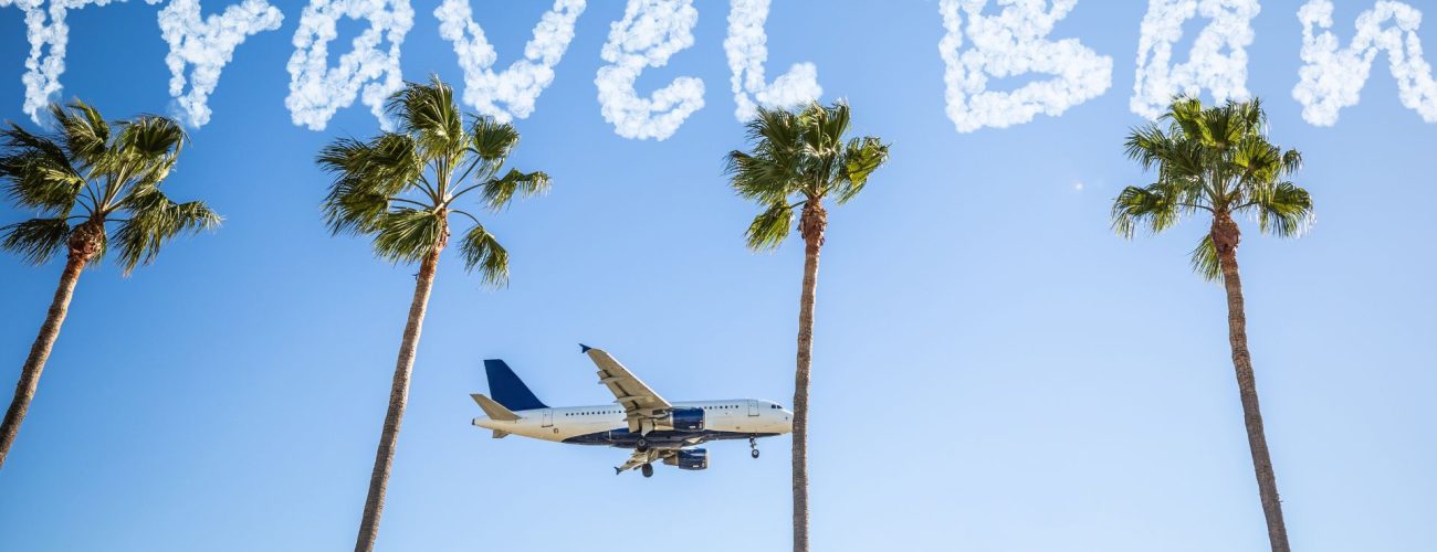 A commercial airplane flying low over four palm trees, with the words 'Travel Ban' written in the sky with cloud-like letters against a clear blue background.