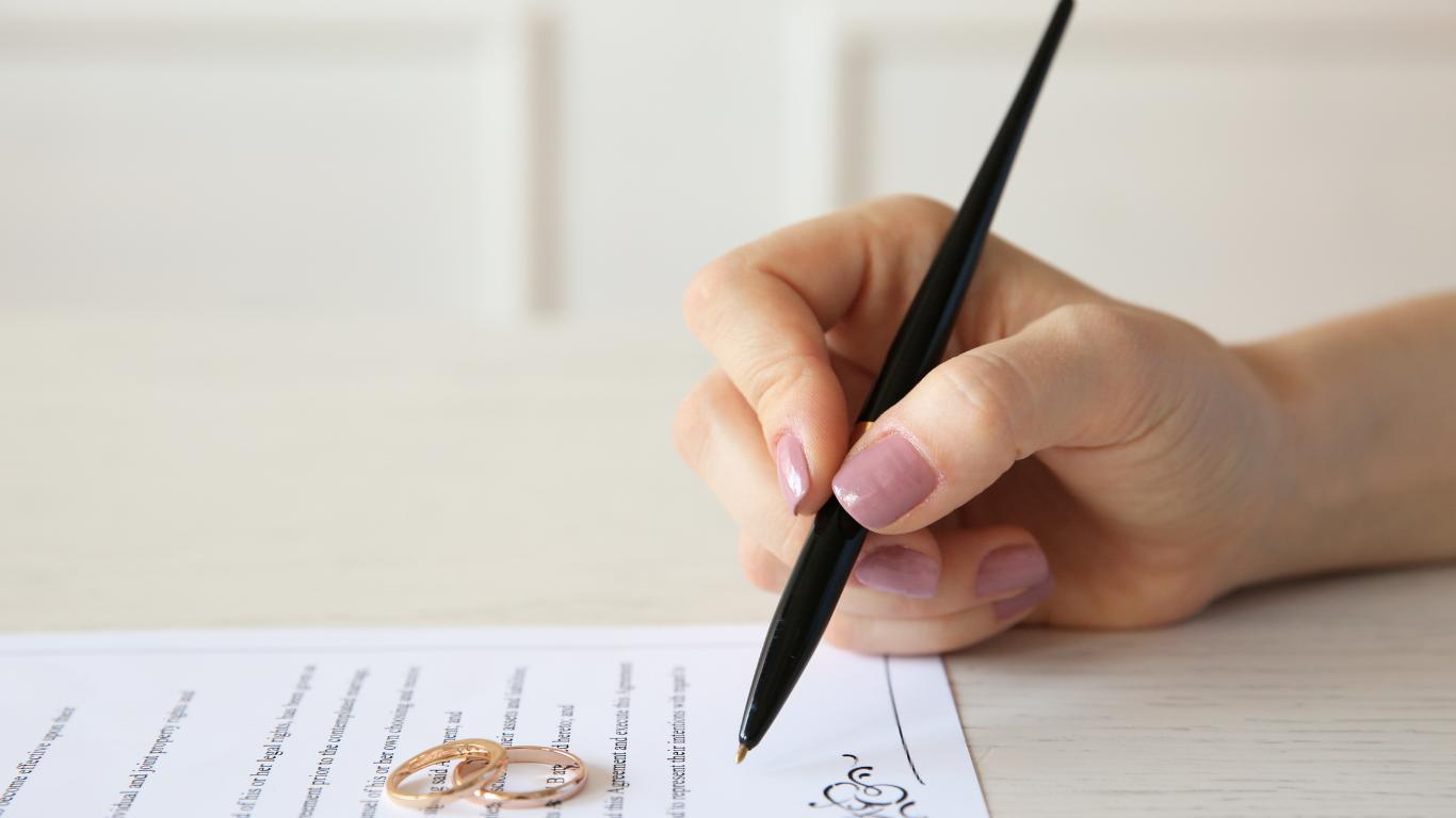 A person's hand is pictured signing a document, likely a marriage certificate, with a pair of gold wedding bands in the foreground, symbolizing the legal and ceremonial union being formalized on paper.