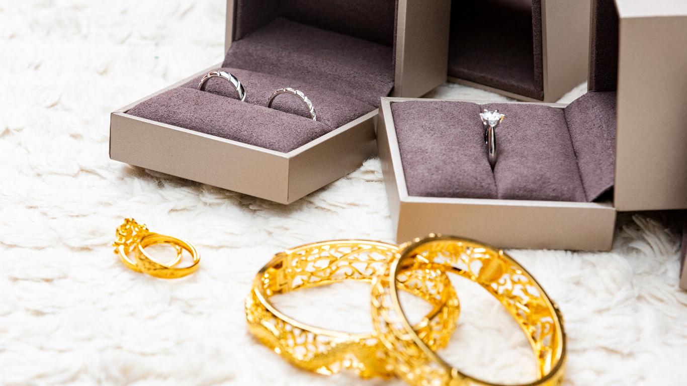 Two open jewelry boxes revealing wedding rings—one with two silver bands and the other with a single solitaire diamond ring—sit atop a plush white surface, with elegant gold bangles resting beside them, together composing a luxurious wedding jewelry set.