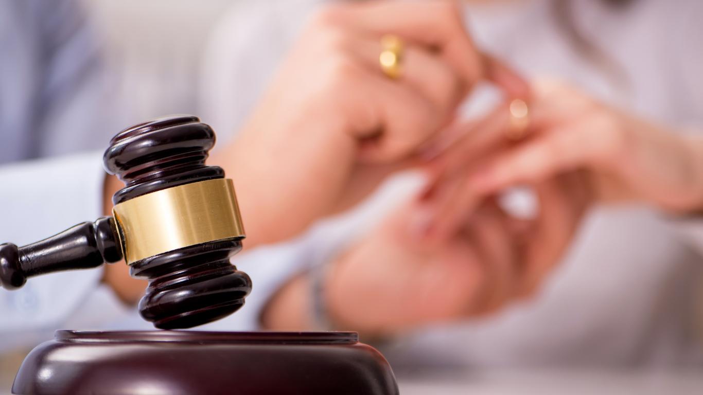 The image focuses on a judge's gavel beside the hands of a person, who appears to be removing or placing a ring on their finger, signifying a legal aspect of marriage, possibly a civil union. The blurred background emphasizes the legal instrument and the act being performed.
