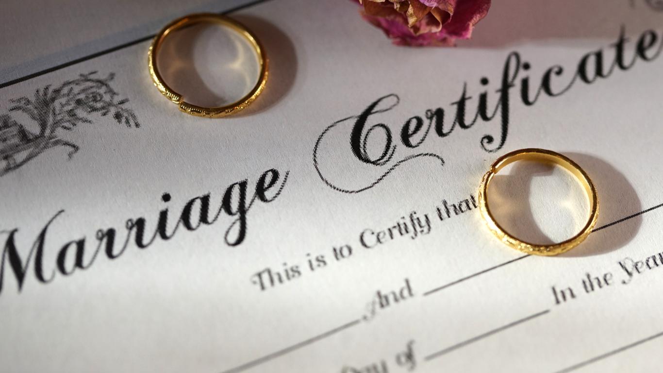 A marriage certificate is adorned with two golden wedding bands, a dried flower adding a touch of romance, all under warm light that casts a soft glow on these symbols of a loving union formalized in matrimony.