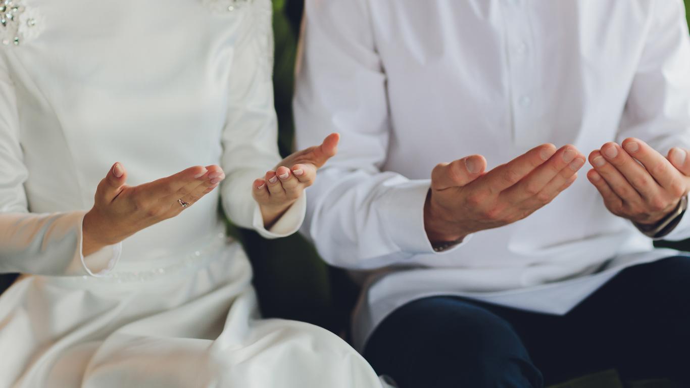 A bride and groom are captured in a moment of devotion, their hands gently raised in prayer or supplication, a gesture indicative of their spiritual commitment and the solemnity of their wedding ceremony. The bride's delicate wedding band is visible, adding a subtle symbol of their union.