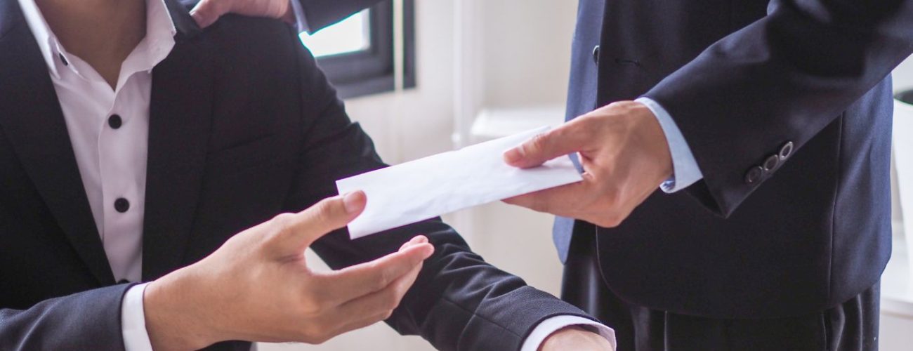 A man in a suit receives an envelope from another person while seated at a desk, with one hand on his shoulder, symbolizing a professional or confidential exchange.