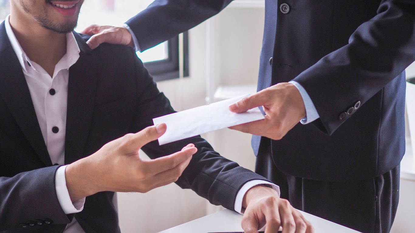 A man in a suit receives an envelope from another person while seated at a desk, with one hand on his shoulder, symbolizing a professional or confidential exchange.