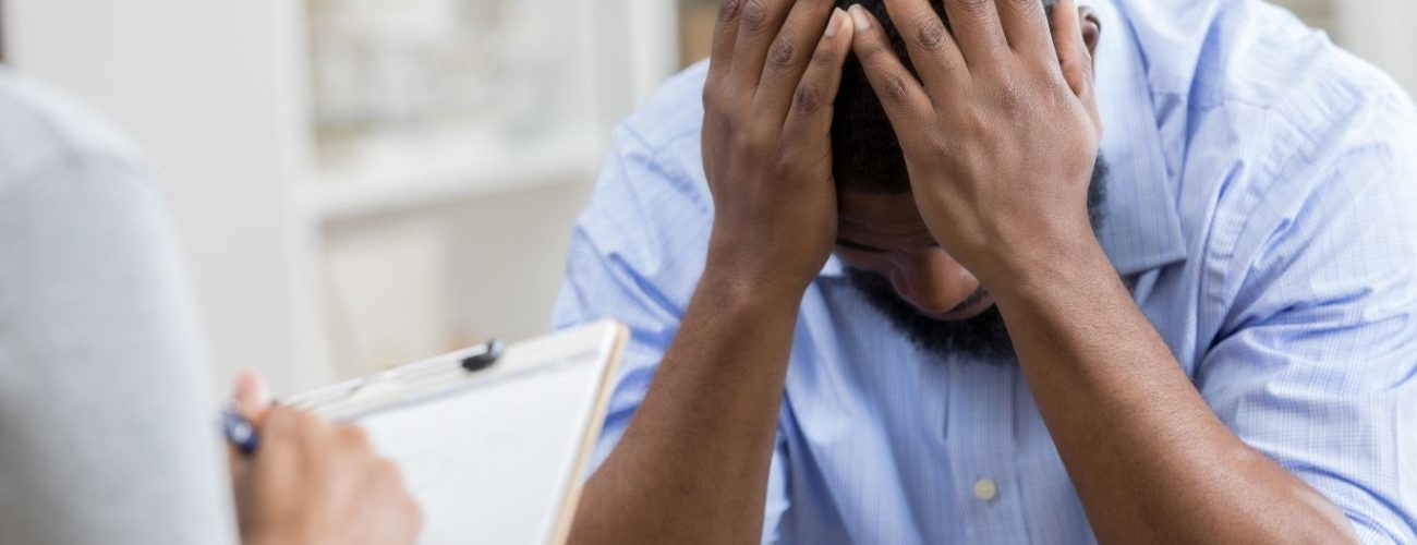 A distressed man sitting at a table during a consultation, holding his head in his hands while a therapist or counselor takes notes.