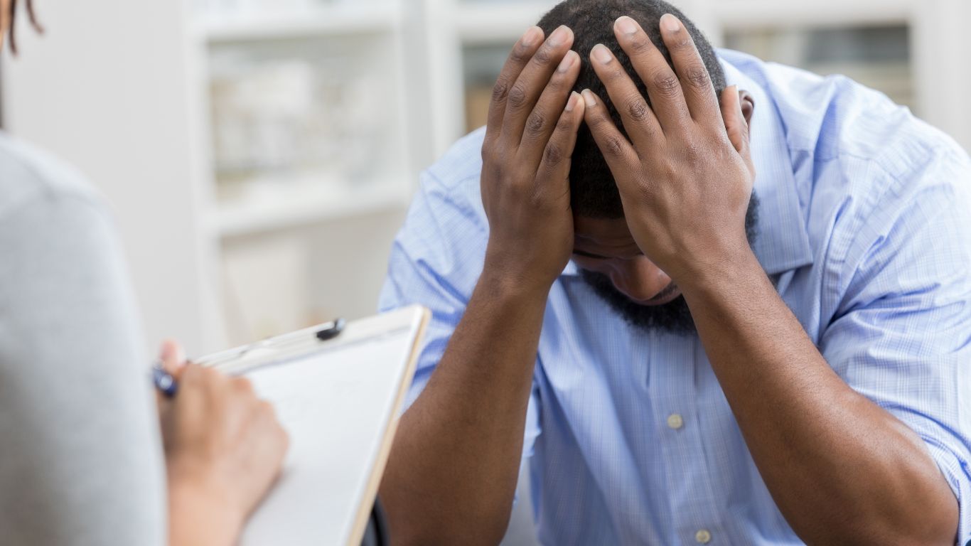 A distressed man sitting at a table during a consultation, holding his head in his hands while a therapist or counselor takes notes.