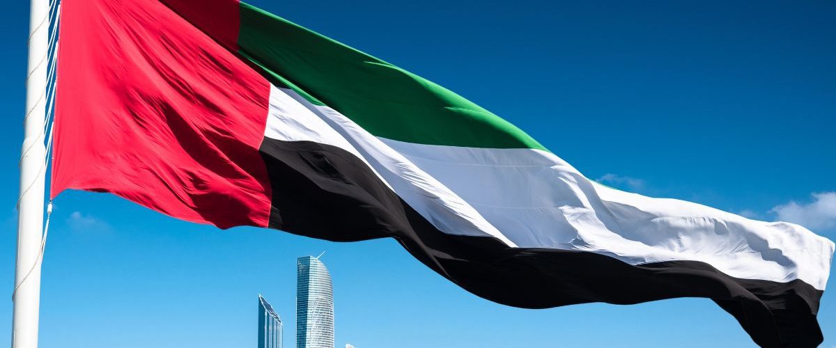 The UAE flag waving prominently in the foreground against a clear blue sky, with modern skyscrapers of Abu Dhabi in the background.