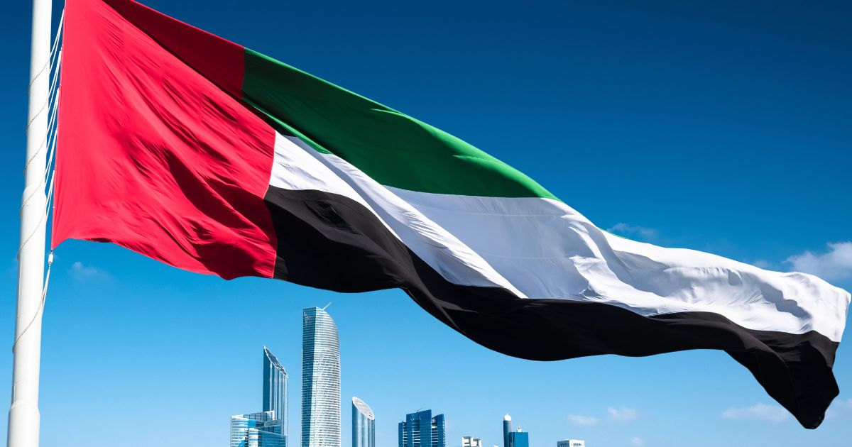 The UAE flag waving prominently in the foreground against a clear blue sky, with modern skyscrapers of Abu Dhabi in the background.