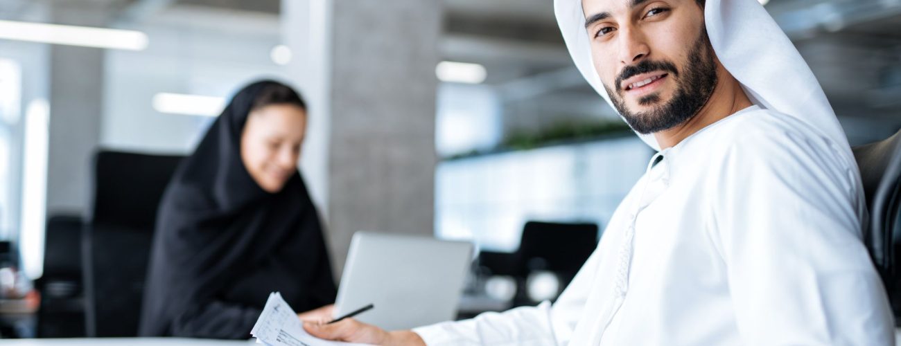 A confident Emirati man dressed in traditional white kandura sitting at a desk, holding documents and looking at the camera with a slight smile. In the background, a woman in a black abaya is focused on her work, using a laptop in a modern office setting. The image represents professional Emirati workers in a business environment, aligning with themes of Emiratisation and workforce participation in the UAE.