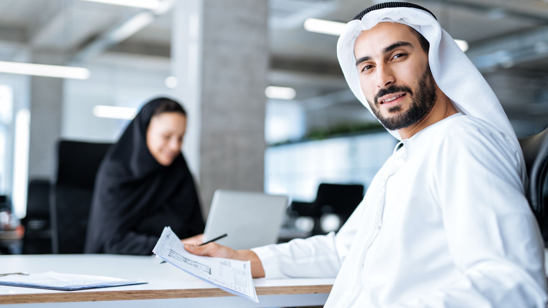 A confident Emirati man dressed in traditional white kandura sitting at a desk, holding documents and looking at the camera with a slight smile. In the background, a woman in a black abaya is focused on her work, using a laptop in a modern office setting. The image represents professional Emirati workers in a business environment, aligning with themes of Emiratisation and workforce participation in the UAE.