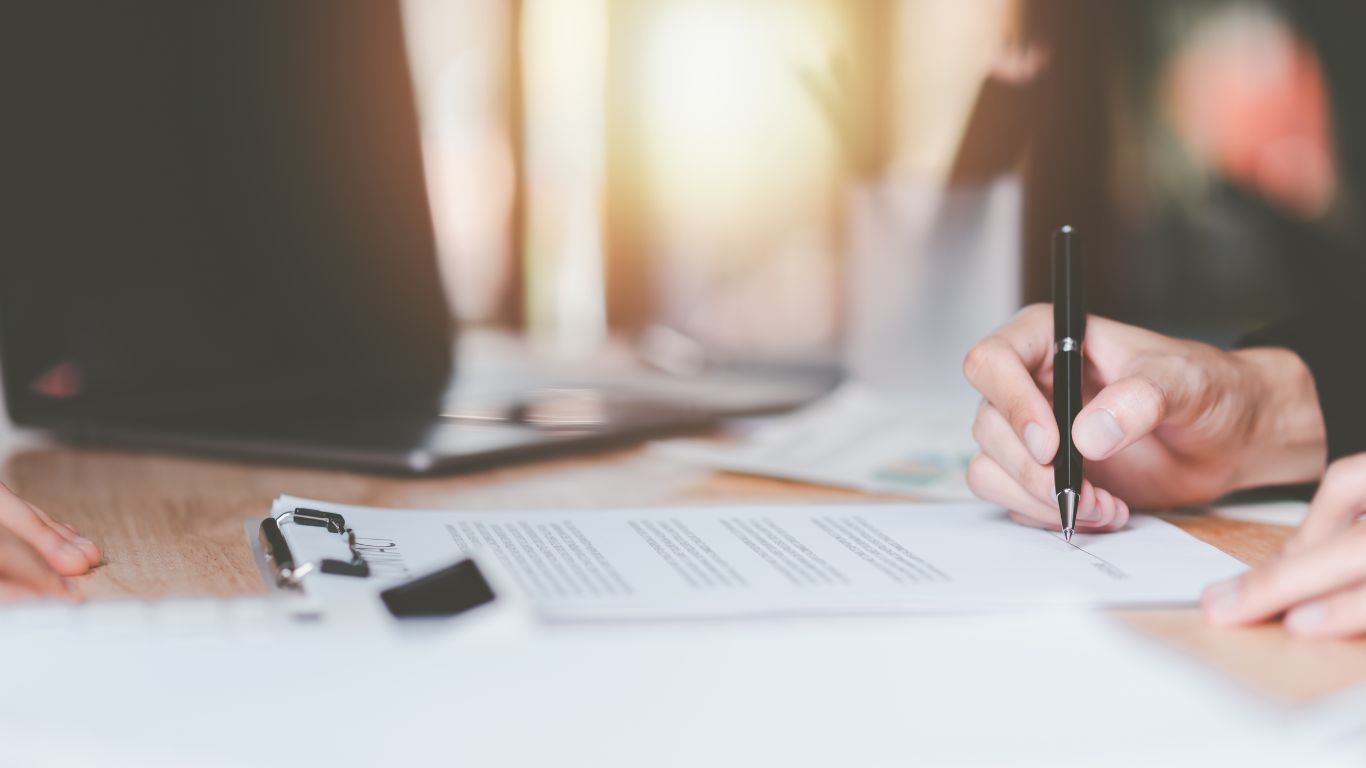 A close-up of hands holding a contract, with one person signing the document. A pen is poised over the signature line, symbolizing the formal agreement or breach documentation process.