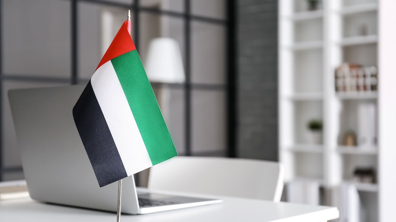 A UAE flag displayed on a small stand beside a laptop in a modern, minimalist office setting, representing national pride or business operations within the UAE, possibly related to UAE visa amnesty.